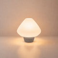 Abajur de mesa Lotus aceso, com cúpula branca plissada e base cinza claro