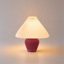 Abajur de mesa Vitta aceso, com cúpula branca plissada e base vermelho marsala