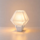 Abajur de mesa aceso, com cúpula branca hexagonal plissada e base branca