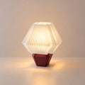 Abajur de mesa aceso, com cúpula branca hexagonal plissada e base vermelho marsala